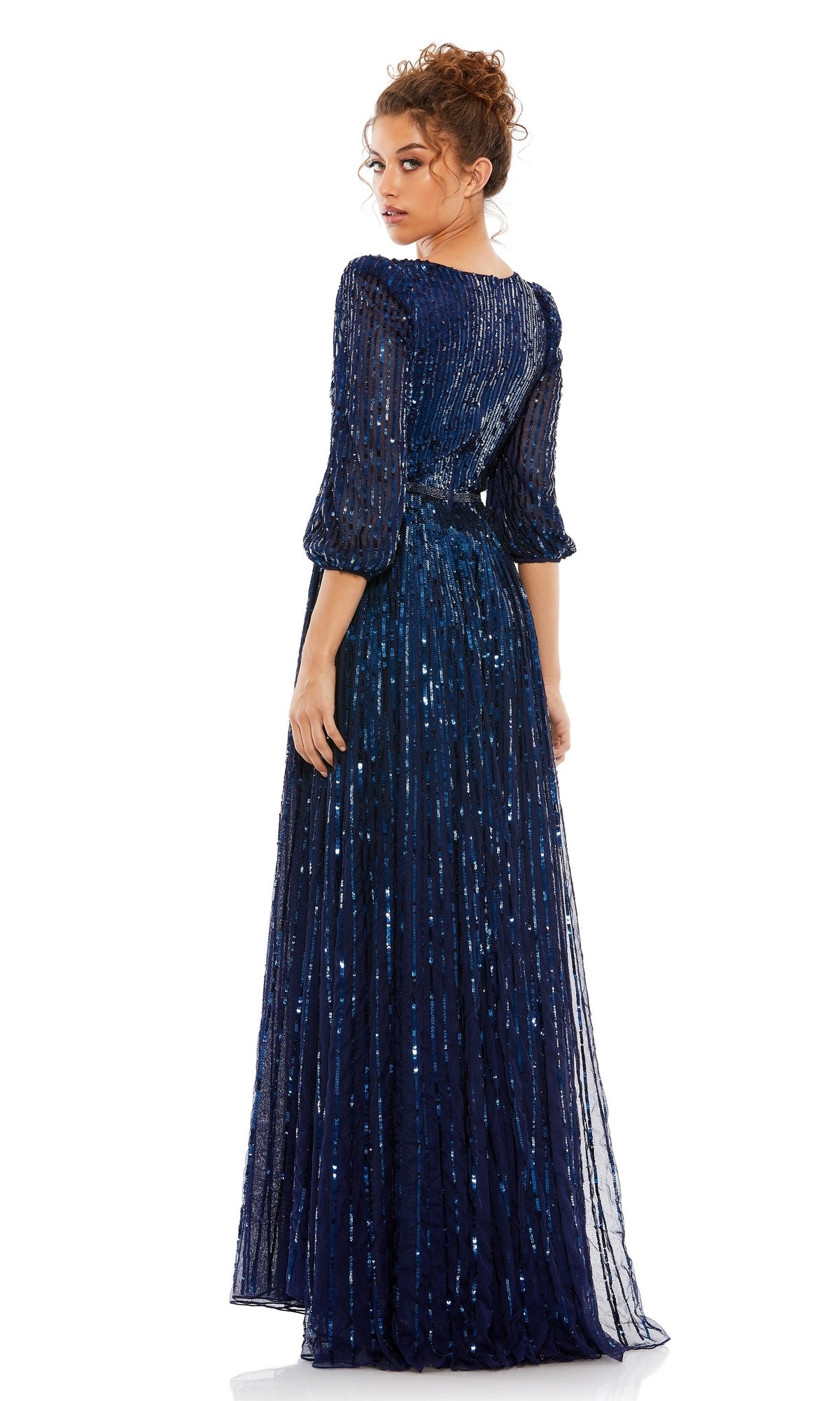 Woman in a floor-length Mac Duggal 5509 long sleeve formal dress with sequins, sheer sleeves, and an open back, glancing over her shoulder against a plain white background.
